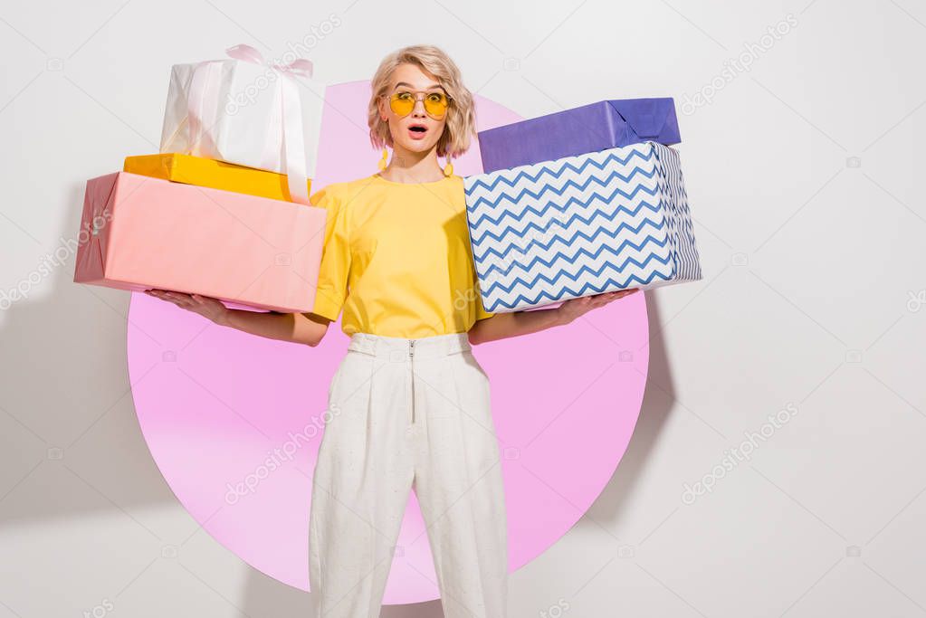 surprised stylish girl holding gift boxes and looking at camera on white with pink circle