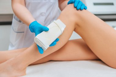 Cropped view of cosmetologist in rubber gloves applying wax on leg clipart