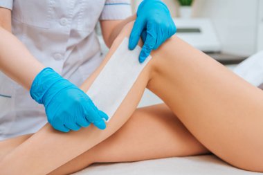 Partial view of cosmetologist in rubber gloves doing leg wax depilation clipart