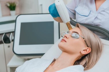 Blonde girl in protective goggles receiving laser treatment in clinic clipart