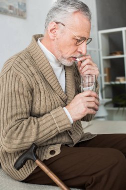 retired man taking pill while sitting on sofa near walking cane 