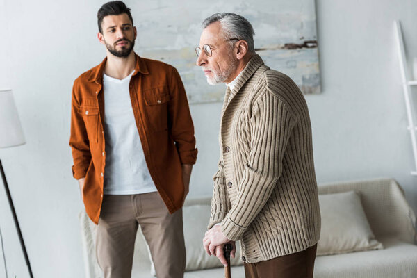 handsome bearded son standing with hands in pockets and looking at sad senior father in glasses