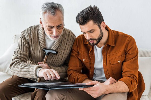 senior father in glasses pointing with finger at photo album near son