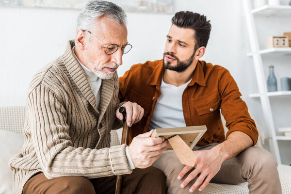 handsome man looking at sad senior dad holding wooden photo frame