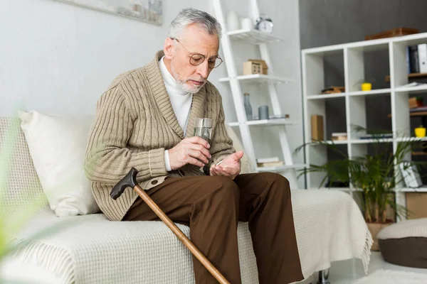 退休男子拿着药丸和一杯水 而坐在沙发上走甘蔗 — 图库照片