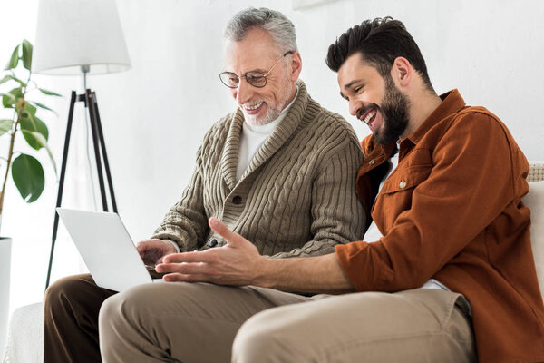 веселый бородатый мужчина жестикулирует, сидя со старшим отцом и глядя на ноутбук
 