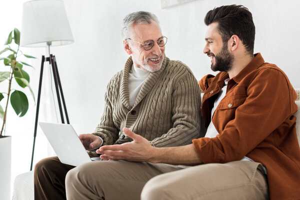 happy bearded man gesturing while sitting with senior father and looking at laptop 