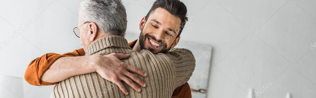 panoramic shot of happy bearded man smiling while hugging elder father at home 