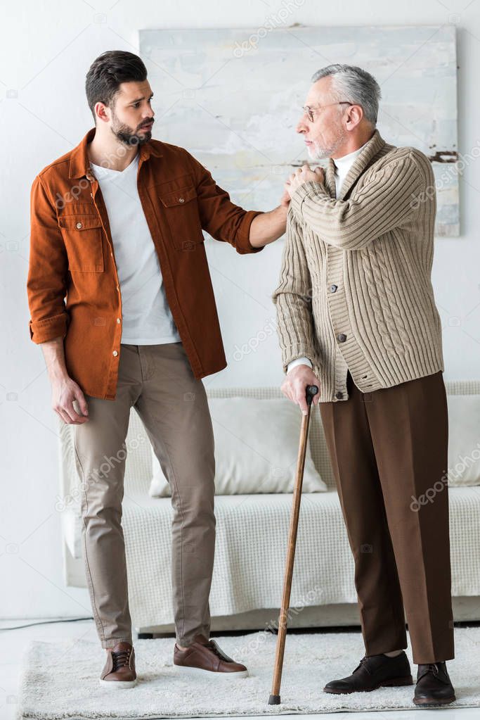 handsome bearded son putting hand on shoulder of sad senior father in glasses with walking stick