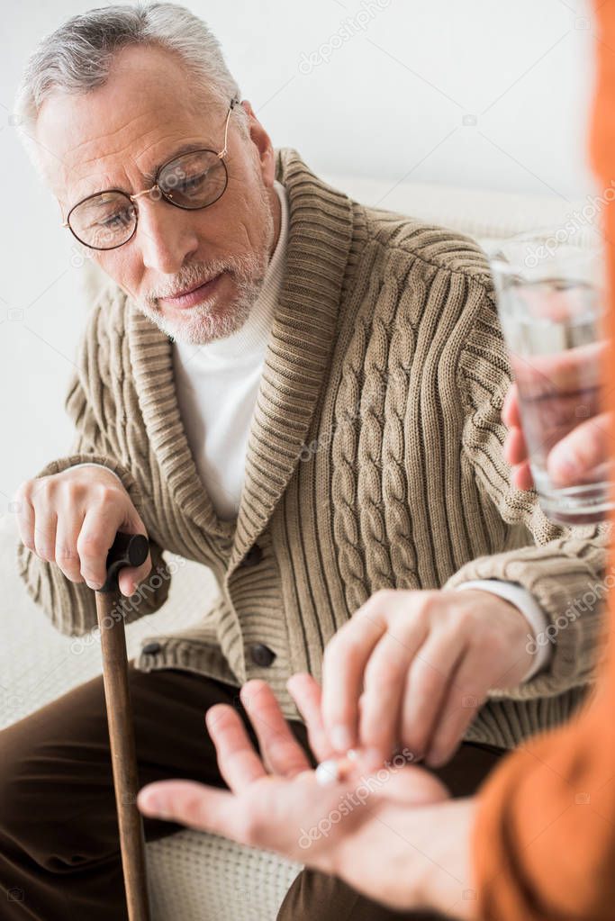 selective focus of retired father taking pills from hand of son holding glass of water 