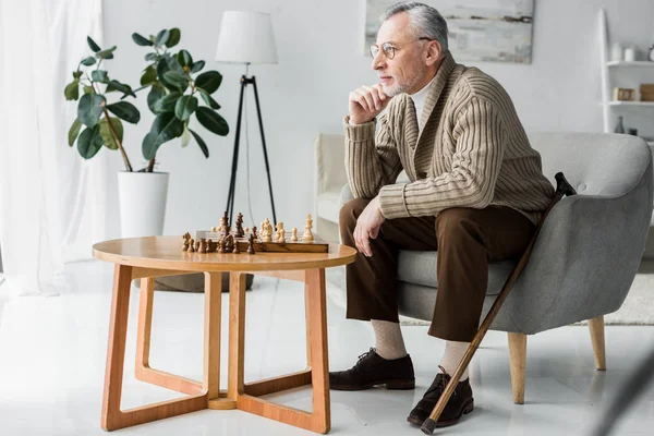 Старший Человек Очках Думает Сидя Рядом Шахматной Доской Дома — стоковое фото