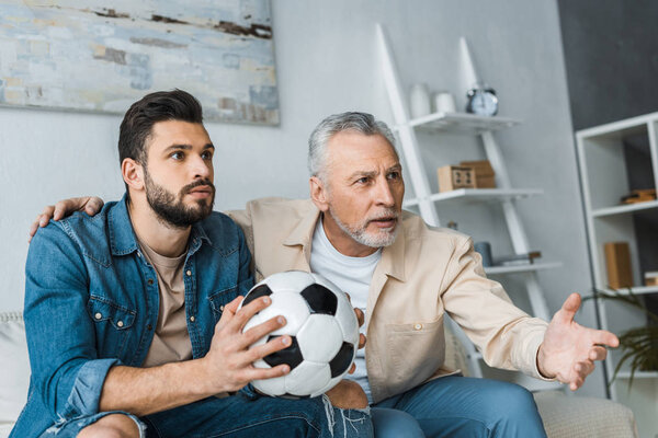 retired man gesturing while watching championship with handsome son holding football 