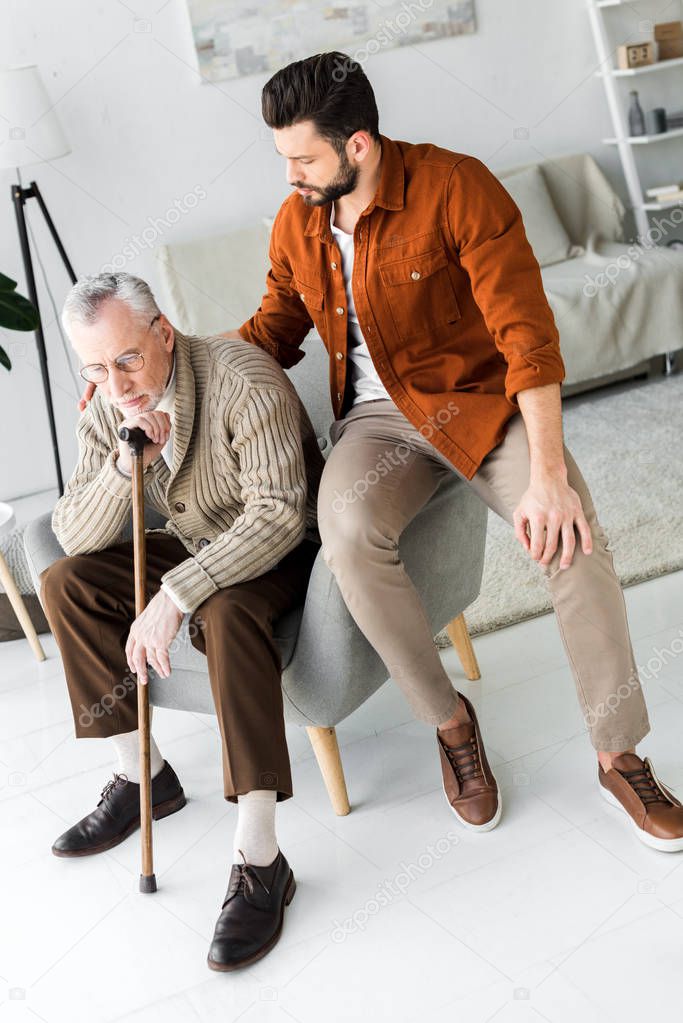 handsome bearded son looking at senior father sitting in armchair at home 