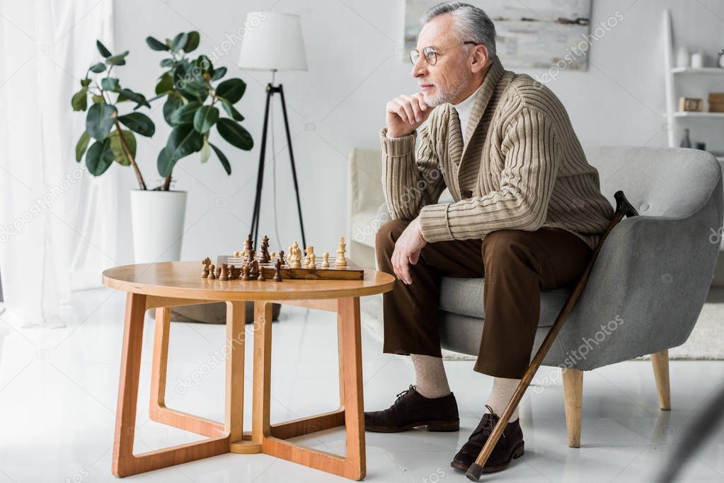 senior man in glasses thinking while sitting near chess board at home