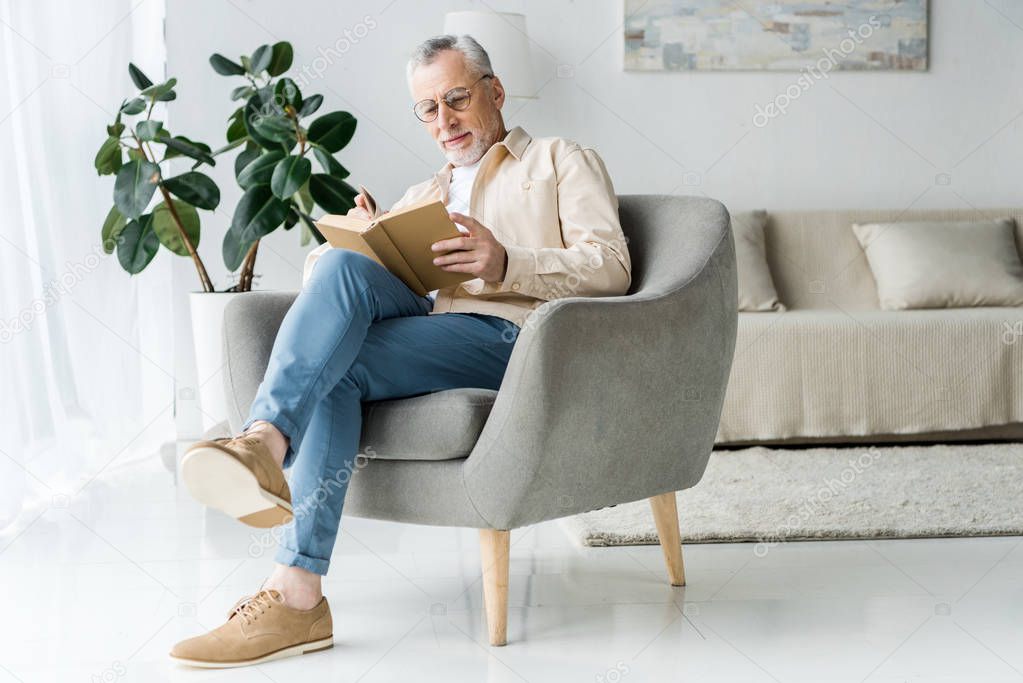 senior man in glasses reading book while sitting in armchair at home 