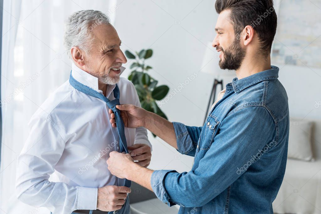 cheerful handsome son helping elegant retired father tying blue tie at home 