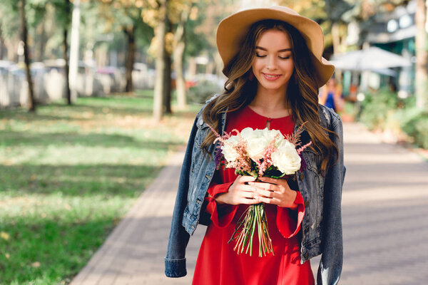 attractive girl in hat looking at flowers while standing in park 