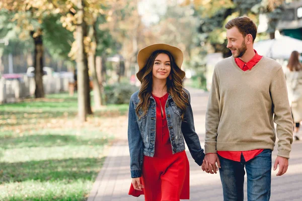 公園で手を握りながら帽子で幸せなガールフレンドを見ている陽気な彼氏 — ストック写真