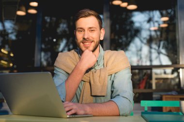 Laptop yakınında oturan neşeli ve yakışıklı Freelancer ve kamera bakıyor 