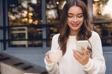 Kupa tutarken akıllı telefona bakarak neşeli genç kadın 