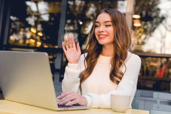 attractive and happy woman waving hand while having video call on laptop