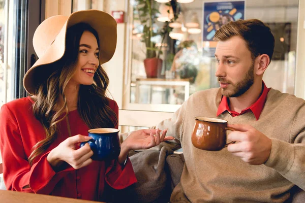 コーヒーのカップを保持しながら男を見ている帽子で幸せな若い女性 — ストック写真