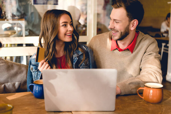 счастливая молодая женщина, держащая кредитную карту и глядя на веселого мужчину возле ноутбука
 