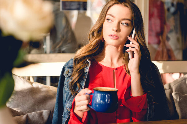 attractive young woman holding cup of coffee and talking on smartphone in cafe 