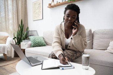 Saçları örgülü genç siyahi bir kadın modern dairesindeki bir telefon görüşmesine not alıyor..