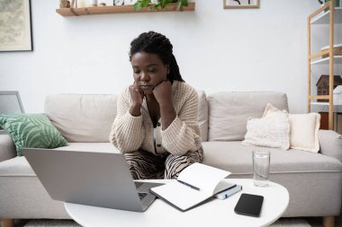 Örgülü saçlı Afro-Amerikalı bir kadın kanepeye oturmuş laptopuna konsantre oluyor..