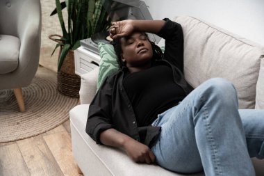 Saçları örgülü bir kadın kanepeye uzanmış, üzgün ve yaşam alanında bunalmış hissediyor..