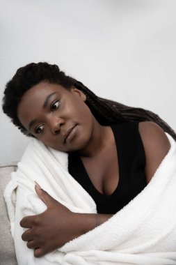 A woman sits quietly at home, wrapped in a blanket, reflecting on her feelings of sadness.