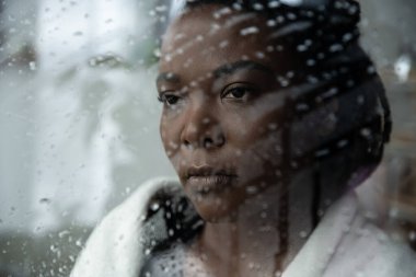 Saçları örgülü bir kadın yağmurlu bir pencereden bakar, evinde derin bir üzüntü hisseder..