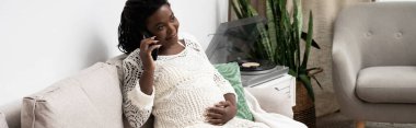 A pregnant woman relaxes on a couch, smiling while holding her belly and talking on the phone, banner