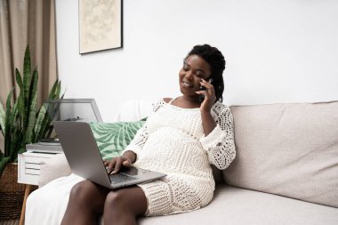 Hamile bir kadın oturma odasında rahat bir şekilde dizüstü bilgisayarda çalışıyor ve telefonda sohbet ediyor..
