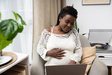 Saçları örgülü hamile bir Afro-Amerikalı kadın evde sessiz vakit geçirip gülümsüyor..
