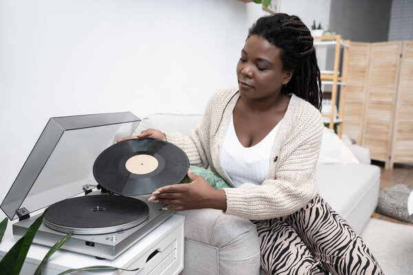 An African American woman with braided hair listens to vinyl records in her cozy living room.