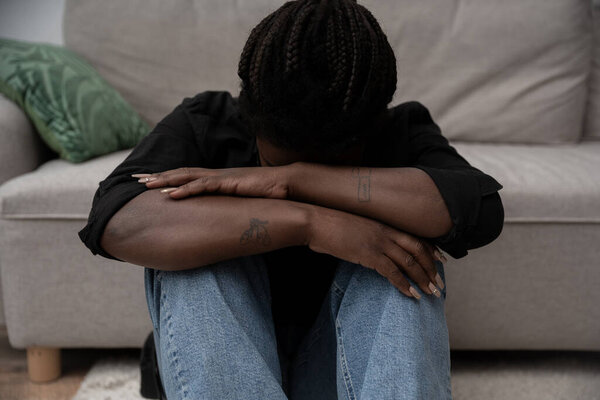 Женщина с плетеными волосами сидит на полу, обнимая колени в момент грусти дома.