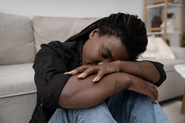 A woman with braided hair sits on the couch, feeling deeply sad and overwhelmed at home.