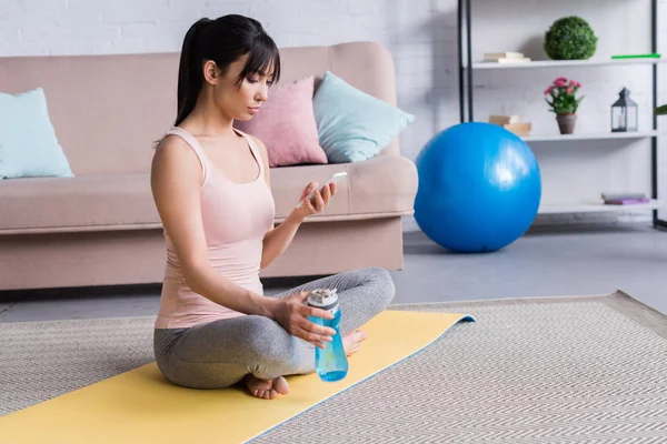 Bella giovane donna rilassante sul tappetino yoga dopo l'allenamento e utilizzando lo smartphone a casa — Foto stock