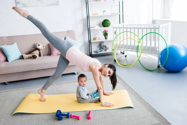 Junge Mutter praktiziert Yoga in einbeiniger, nach unten gerichteter Hundehaltung, während ihr kleines Kind zu Hause auf Matte sitzt — Stockfoto