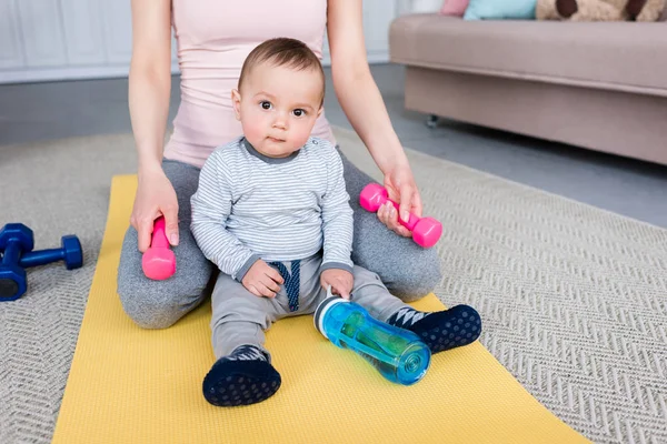 Обрезанный снимок матери с гантелями и маленький ребенок сидит на коврике для йоги дома — стоковое фото