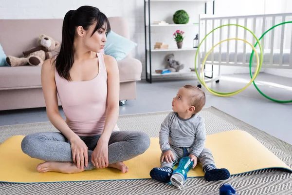 Молодая красивая мать и маленький ребенок сидят на коврике для йоги дома и смотрят друг на друга — стоковое фото