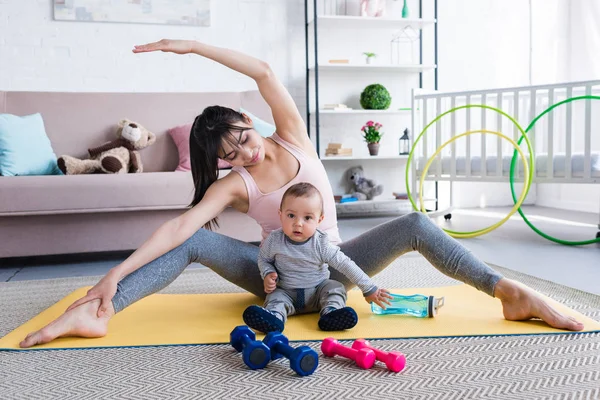 Junge sportliche Mutter dehnt sich, während sie mit Kind auf Matte sitzt — Stockfoto