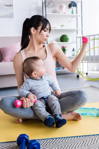 Joven madre deportista haciendo ejercicio con mancuernas mientras se sienta en la estera con el niño - foto de stock