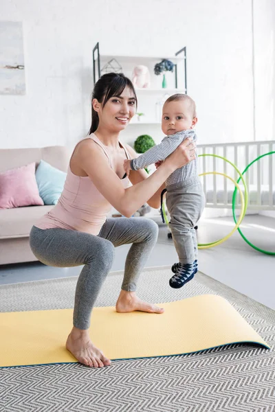 Young sportive mother carrying her little child while doing squats — Stock Photo