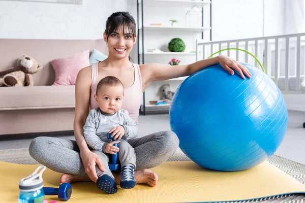Jeune mère souriante et petit enfant assis sur un tapis de yoga à la maison avec balle en forme — Photo de stock