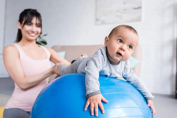 Young happy mother playing with little child while he lying on fit ball at home — Stock Photo