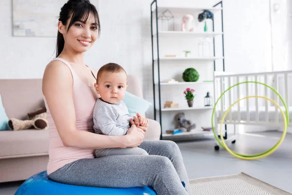 Young smiling mother sitting on fit ball with little child at home — Stock Photo