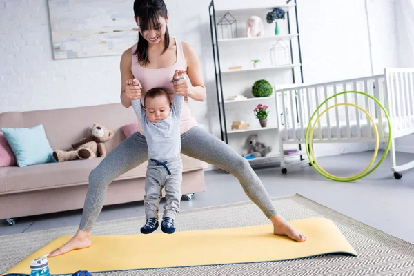 Joven madre deportiva llevando a un niño pequeño mientras está de pie en la esterilla de yoga en casa - foto de stock